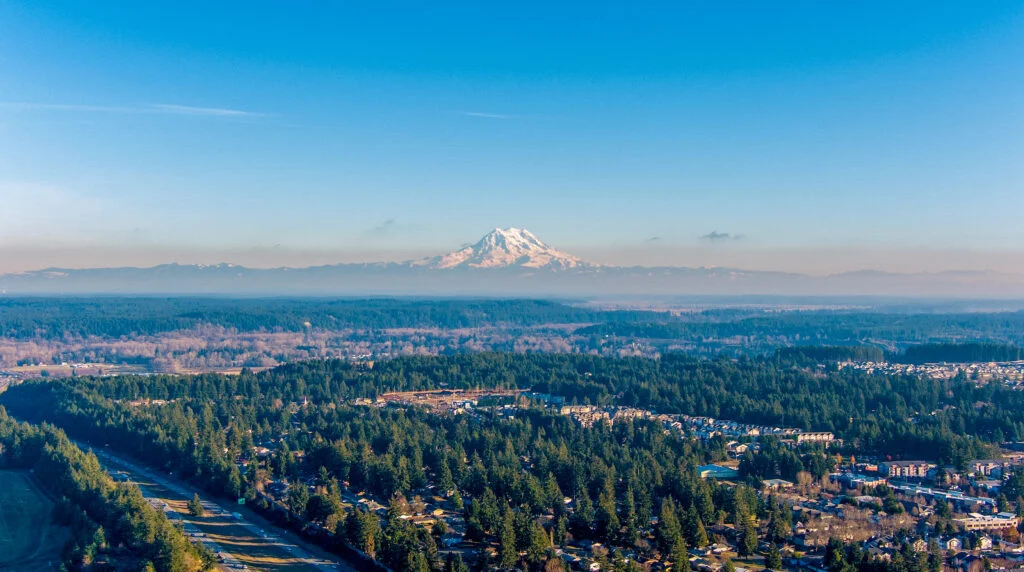 An aerial photo of Lacey Washington with neighborhoods and trees in the foreground and Mt Rainier in the background. Lacey is a city near JBLM