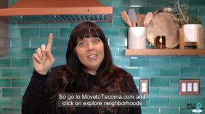 A woman with long brown hair and a black blazer gestures while speaking and standing in a kitchen talking about MovetoTacoma.com