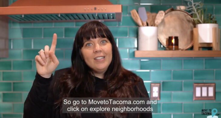 A woman with long brown hair and a black blazer gestures while speaking and standing in a kitchen talking about MovetoTacoma.com