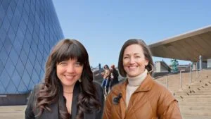 A screenshot of two people one with long dark hair one with short brown hair standing in front of the glass museum in downtown tacoma washington.