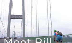 A picture of bill in a bike helmet, tie died shirt,and blue shorts biking over the narrows bridge. A photo from his interview about what he likes about living in gig harbor wa