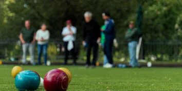 Three lawn bowling balls in the foreground with people from the Tacoma Lawn Bowling Club bowling across a green lawn in the distance.