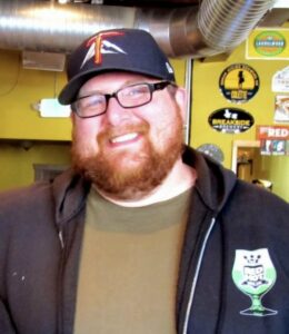Chris Miller owner of the red hot in Tacoma's 6th ave neighborhood. He wears a hat, a green shirt, a black hoodie, and has a red beard.