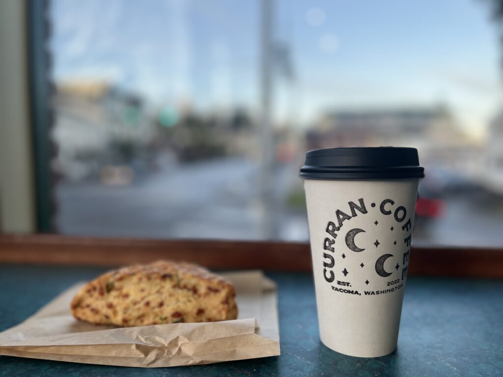 a photo of a cup of coffee to go with the words, "Curran Coffee est 2022 Tacoma, Washington" sits on the counter next to a cheddar scone on a napkin overlooking the old town neighborhood of tacoma. 