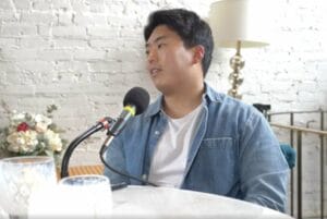 Kevin Lee wears a white tshirt and sits and a table with a podcasting microphone in Tacoma, Washington wearing a white t and a denim shirt