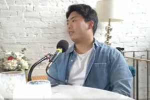 Kevin Lee wears a white tshirt and sits and a table with a podcasting microphone in Tacoma, Washington wearing a white t and a denim shirt