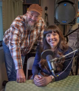 Doug Mackey, a person in a hat and flannel shirt with a long gray beard stands next to Marguerite Martin, a person with long brown hair and a blue shirt in front of a podcast mic where they record the Move to Tacoma Podcast in Tacoma Washington.