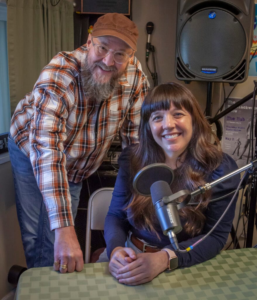 Doug Mackey, a person in a hat and flannel shirt with a long gray beard stands next to Marguerite Martin, a person with long brown hair and a blue shirt in front of a podcast mic where they record the Move to Tacoma Podcast in Tacoma Washington.