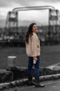Marguerite Martin Tacoma real estate agent and creator of Move to Tacoma stands in front of the Murray Morgan Bridge holding a cup of coffee and wearing a cable knit sweater.