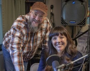 Doug Mackey, a person in a hat and flannel shirt with a long gray beard stands next to Marguerite Martin, a person with long brown hair and a blue shirt in front of a podcast mic where they record the Move to Tacoma Podcast in Tacoma Washington.
