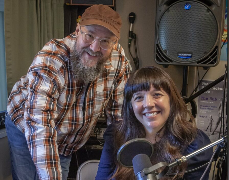 Doug Mackey, a person in a hat and flannel shirt with a long gray beard stands next to Marguerite Martin, a person with long brown hair and a blue shirt in front of a podcast mic where they record the Move to Tacoma Podcast in Tacoma Washington.
