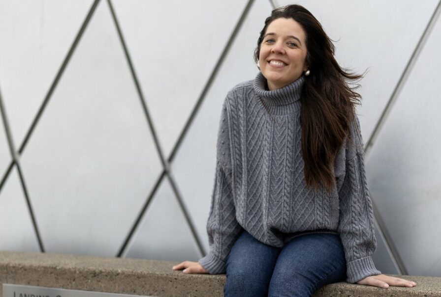 Tacoma real estate agent Marguerite Martin, a person with long brown hair wearing a grey sweater sits in front of the mirrored surface of the Tacoma Glass Museum.