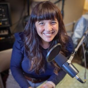 Marguerite Martin recording the move to Tacoma podcast. A person with long brown hair and a blue shirt sits at a table with a podcast microphone in Tacoma Washington.