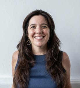 a photo of marguerite martin tacoma real estate agent smiling on a grey background with long dark hair and a blue blouse