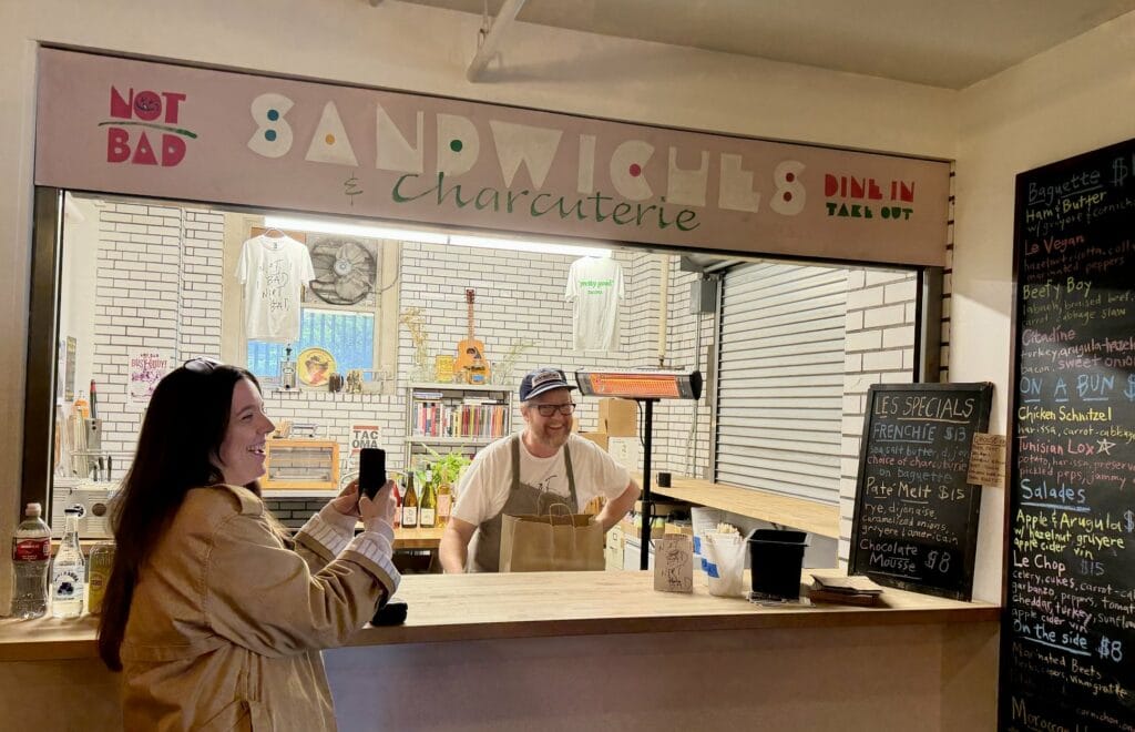 Marguerite Martin wears a long trench coat and holds up her phone while Not Bad Sandwiches owner Dave laughs