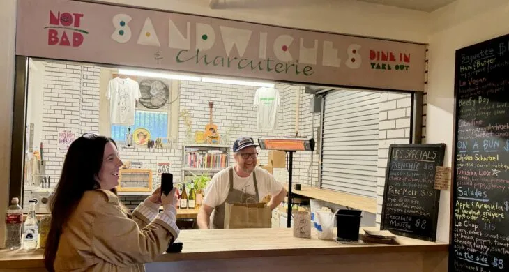 Marguerite Martin wears a long trench coat and holds up her phone while Not Bad Sandwiches owner Dave laughs