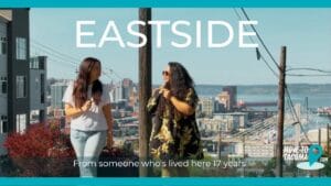 Two tacoma real estate agents with long hair walking up McKinley Hill talking to each other with Downtown Tacoma in the background with the words, "Eastside: From someone whose lived here 17 years"